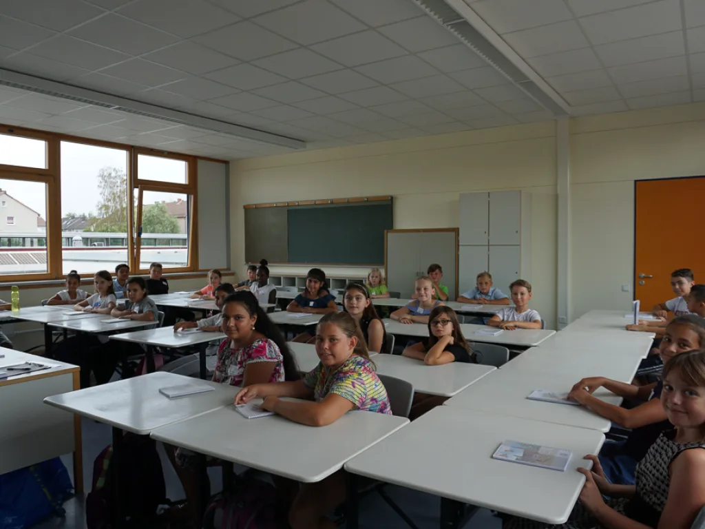 Kinder sitzen an Tischen, im Hintergrund die Tafel.